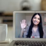 A woman in a video chat on a laptop screen waves to another, shown in a smaller window on the screen. A coffee cup is next to the laptop.