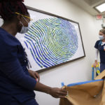 Women in surgical masks and scrubs gather around a framed picture of a fingerprint. Two of the women stand on a ladder and lower a large sheet of paper which had previously been used to cover the image.