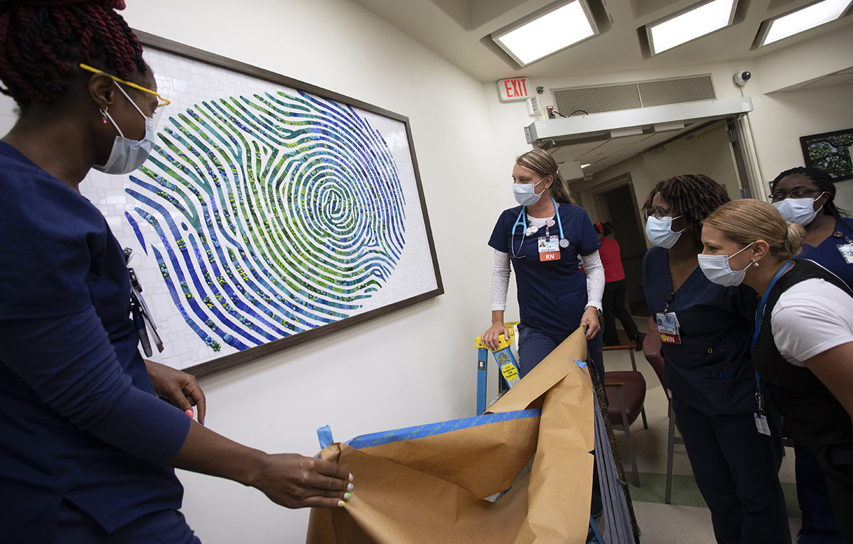 Women in surgical masks and scrubs gather around a framed picture of a fingerprint. Two of the women stand on a ladder and lower a large sheet of paper which had previously been used to cover the image.
