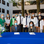 A group of medical students stand behind a table with the Dean of the College of Medicine and another woman seated at a table in front of them. Everyone is outdoors and wearing masks.
