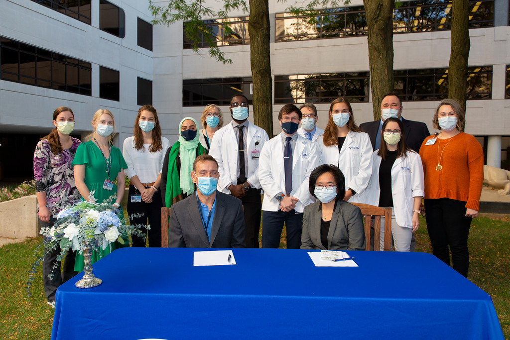 A group of medical students stand behind a table with the Dean of the College of Medicine and another woman seated at a table in front of them. Everyone is outdoors and wearing masks.