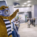 The Penn State Nittany Lion stands in the foreground, with several people in the background cheering as an individual walks through a doorway.
