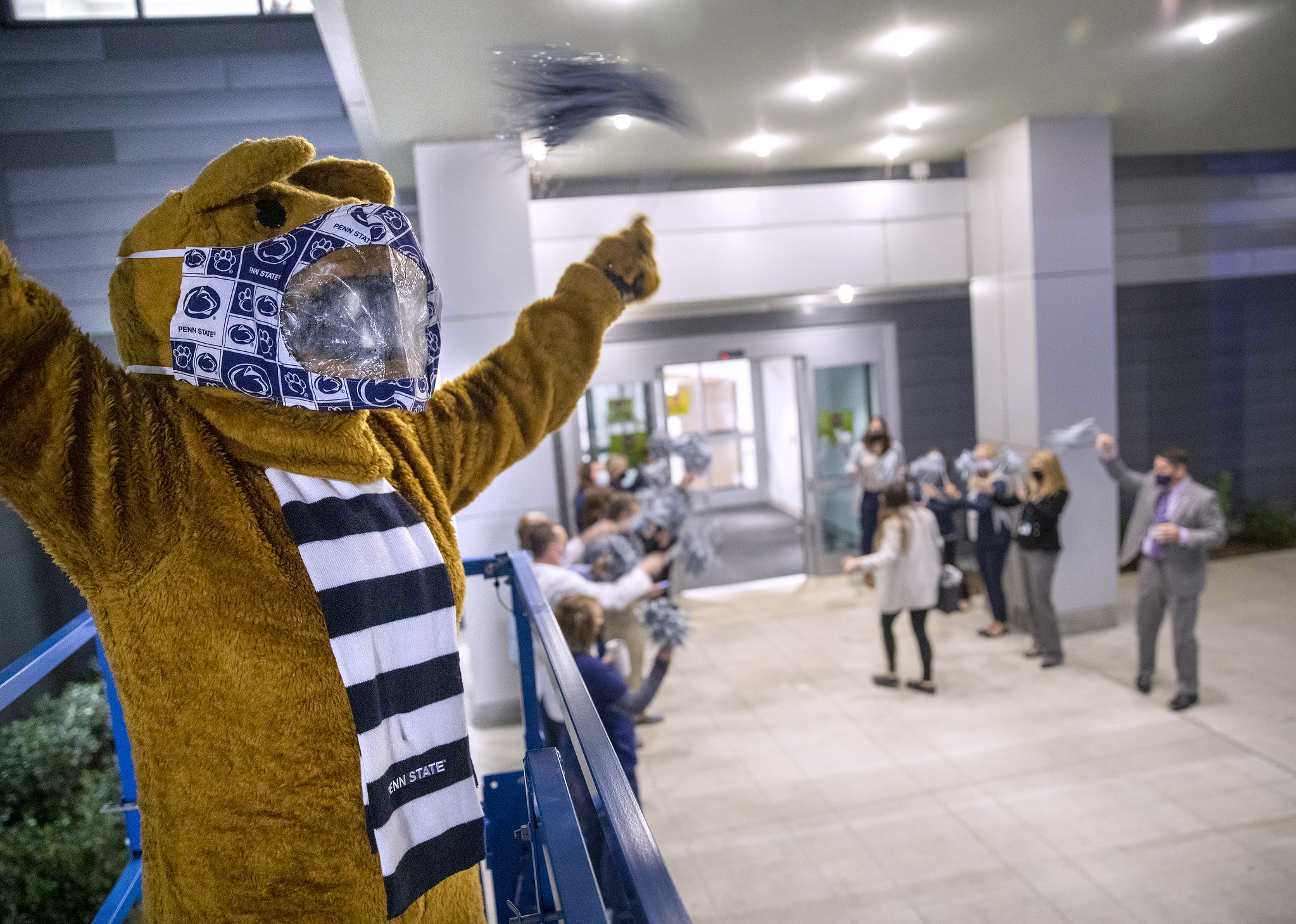 The Penn State Nittany Lion stands in the foreground, with several people in the background cheering as an individual walks through a doorway.