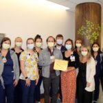 A dozen members of the Pediatric Intermediate Care Unit at Penn State Health Children’s Hospital, wearing masks and scrubs, stand together in a hospital corridor while displaying an award certificate.