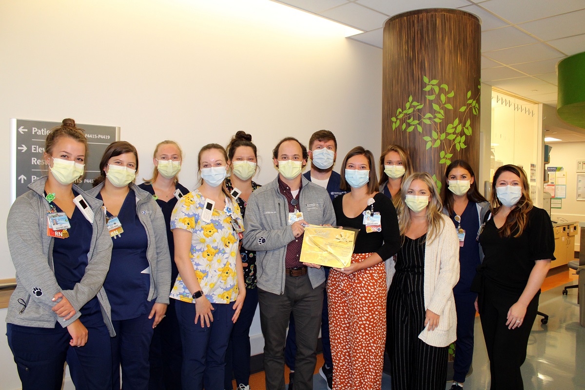 A dozen members of the Pediatric Intermediate Care Unit at Penn State Health Children’s Hospital, wearing masks and scrubs, stand together in a hospital corridor while displaying an award certificate.
