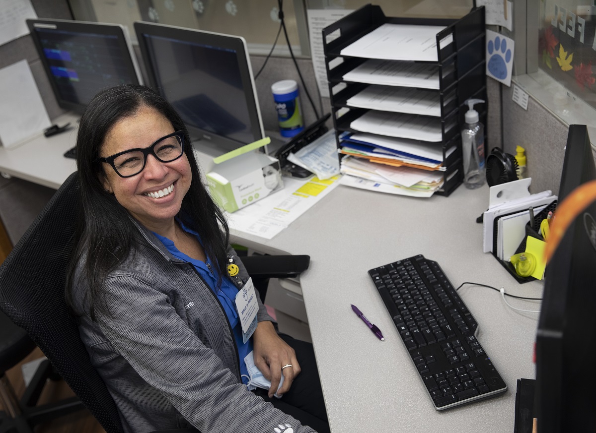 Margarita Biascochea smiles as she sits behind a desk with a computer monitor in front her. She has long hair, glasses and wears a zip-up sweat suit jacket over her shirt.