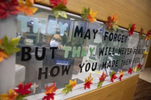 Margarita Biascochea is seen through the glass window in front of her desk area, talking on the phone. The window is decorated with fall leaf stickers and the words “They may forget your name, but they will never forget how you made them feel!”