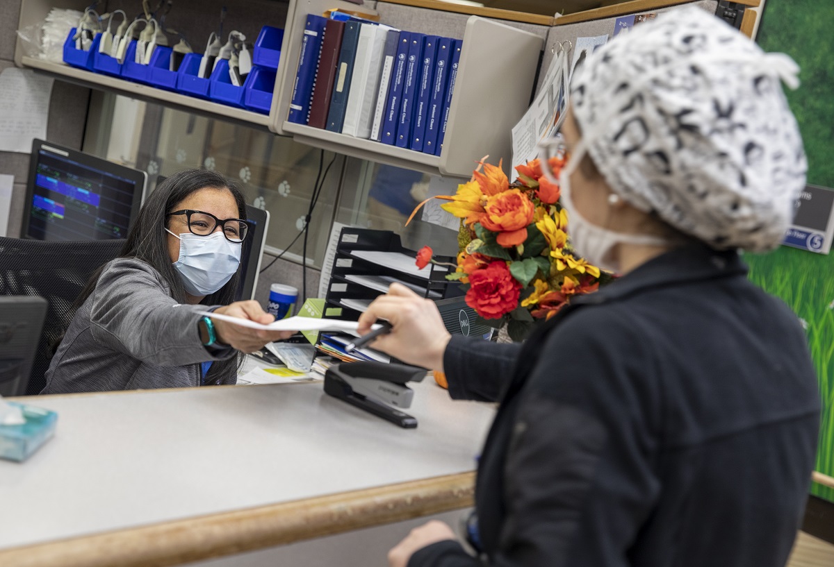 Margarita Biascochea, seated at her desk and wearing a face mask, hands a piece of paper over a counter to a woman who is seen from behind. A stapler and a vase of flowers are on the counter.