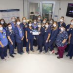 More than 20 staff on the 6th Floor Acute Care Unit, wearing scrubs and face masks, stand in a semi-circle in a hospital hallway. A woman in the middle of the group holds the award certificate in her hand.