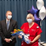 Woman in red uniform wearing a face mask holds bouquet of flowers as man in business suit and mask looks at her and claps.