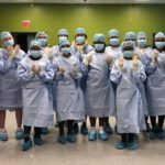Nineteen high school students stand together in a room with a green wall wearing blue surgical gowns, masks and hair and shoe coverings. They are wearing surgical gloves and holding up their hands.