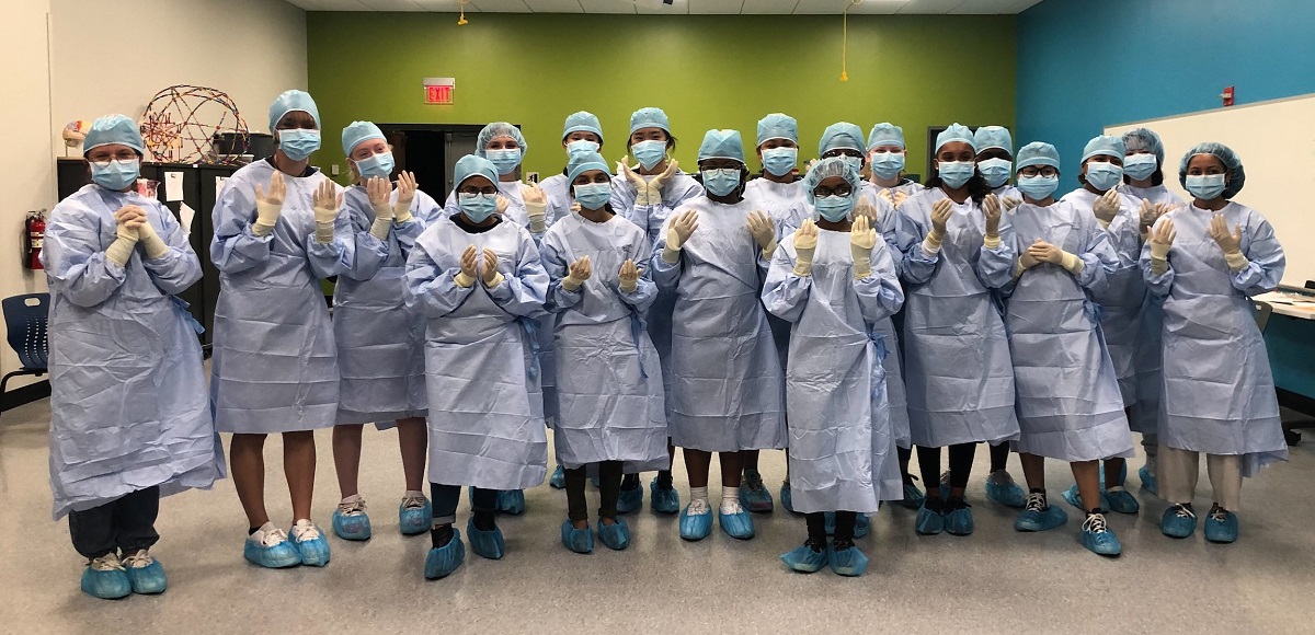 Nineteen high school students stand together in a room with a green wall wearing blue surgical gowns, masks and hair and shoe coverings. They are wearing surgical gloves and holding up their hands.