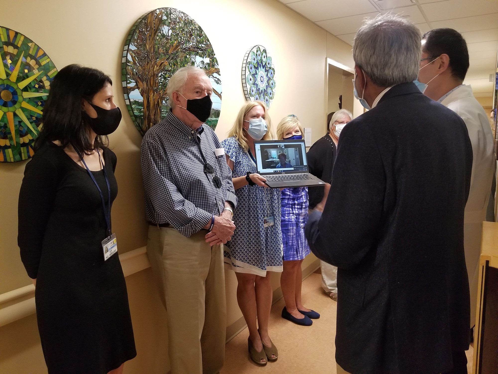 Six people, one holding up a laptop, stand in a hallway with artwork on the wall behind.