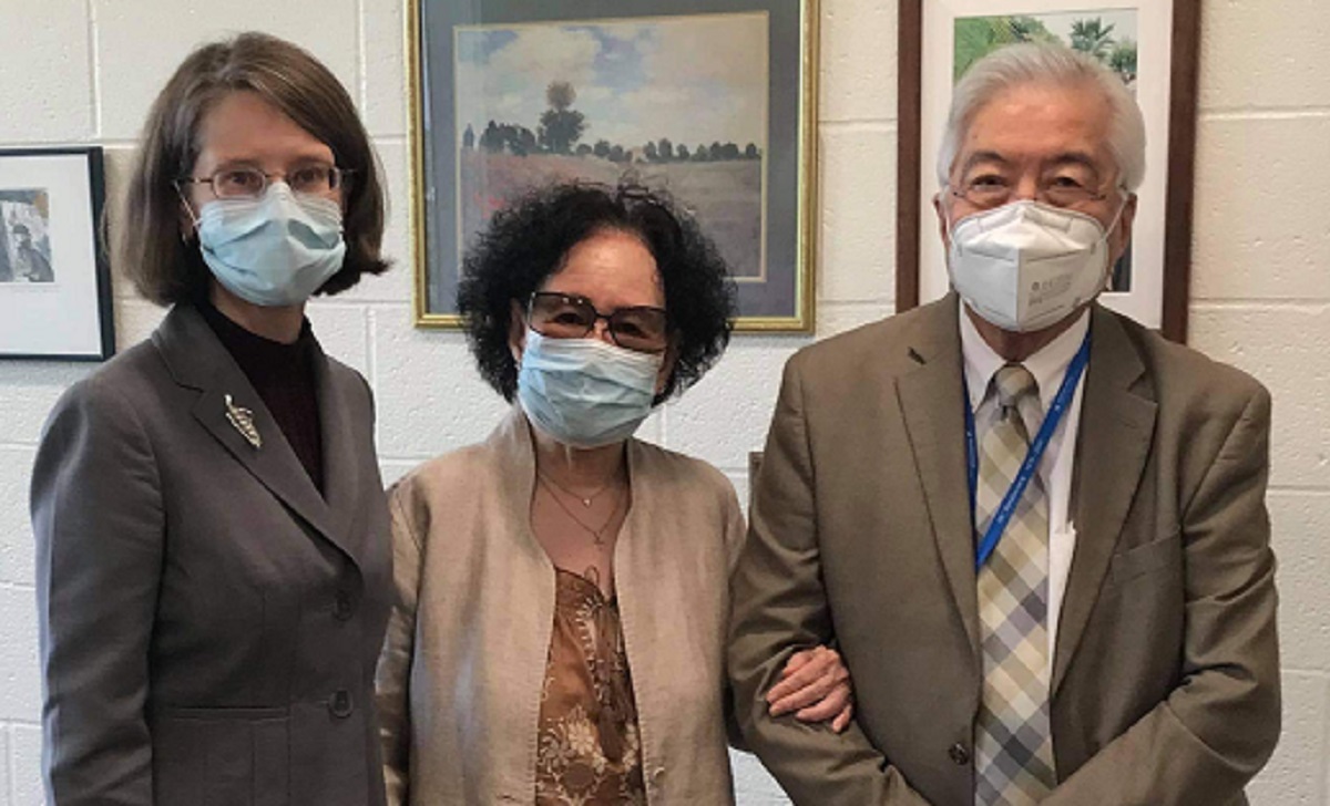 Three people stand next to one another in a room, all wearing face masks. One woman wears a brown suit, and another woman wears a brown jacket. She has her arm linked with a man who is wearing a brown suit and blue checkered tie.