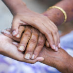 A close-up photo of two sets of adult hands with the one person's hands cradling the other person's hands to convey a sense of support.
