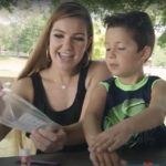 A young mother and her son sit at a picnic table in a park pavilion and work on a craft together.