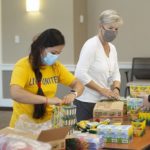 Three women stand at a table and unpack boxes of crayons. The woman on the left has long hair in a brad and is wearing a “Live United T-shirt.” The woman in the middle has short hair and is wear a long-sleeved top. The woman on the right has her hair in a ponytail and is wearing a United Way T-shirt.