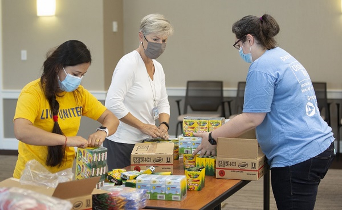 Three women stand at a table and unpack boxes of crayons. The woman on the left has long hair in a brad and is wearing a “Live United T-shirt.” The woman in the middle has short hair and is wear a long-sleeved top. The woman on the right has her hair in a ponytail and is wearing a United Way T-shirt.
