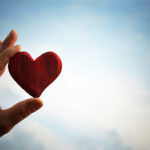 Photo showing a person's hand holding a red, wooden heart against a blue sky.