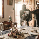 A dinner table is set in the foreground. Just beyond that, a woman lets another woman in the front door, as the two exchange elbow bumps.
