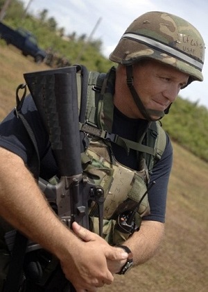 Employee Marvin Smith wears a camouflage vest and helmet while holding a gun pointed toward the ground.