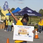 Michelle Phillips of United Way of the Capital Region holds a pom-pom shaker in the air and a sign that reads “Stuff the Bus” for Day of Caring. Behind her are two pop-up tents and people standing in small groups and talking.