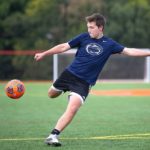Brayden Sunho purses his lips as he gets ready to kick a soccer ball that is in mid-air. Brayden, who is wearing shorts and a shirt with a Penn State Nittany lion logo on the front, is running on a soccer field marked with goal lines.