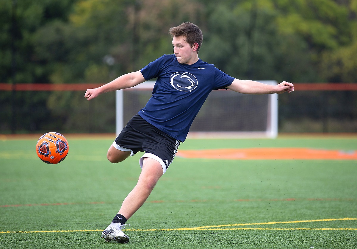 Brayden Sunho purses his lips as he gets ready to kick a soccer ball that is in mid-air. Brayden, who is wearing shorts and a shirt with a Penn State Nittany lion logo on the front, is running on a soccer field marked with goal lines.