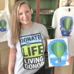 Lauren Gydosh holds the picture she painted of a hot air balloon that says “Donate Life,” surrounded by sky and clouds that contain the words “hope” and “love.” A T-shirt and tote bag bearing the same design hang on cabinet doors behind her. Lauren, who has long, straight hair, is wearing shorts and a T-shirt that says “Donate Life Living Donor.”