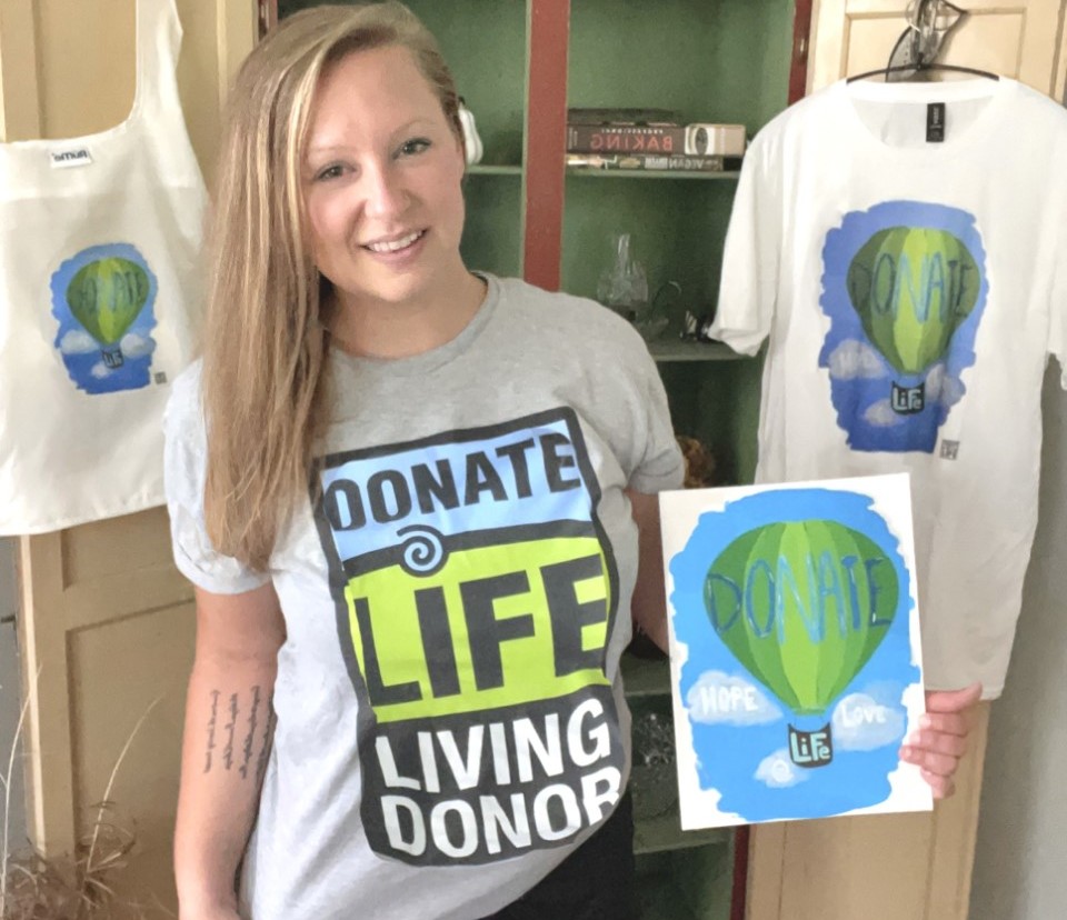 Lauren Gydosh holds the picture she painted of a hot air balloon that says “Donate Life,” surrounded by sky and clouds that contain the words “hope” and “love.” A T-shirt and tote bag bearing the same design hang on cabinet doors behind her. Lauren, who has long, straight hair, is wearing shorts and a T-shirt that says “Donate Life Living Donor.”