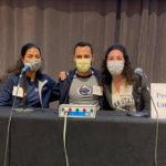Physician Assistant students Jasmin Pimentel, Jonathan McKinney and Maria Gillio sit behind a table wearing face masks, with a sign that says Penn State University.