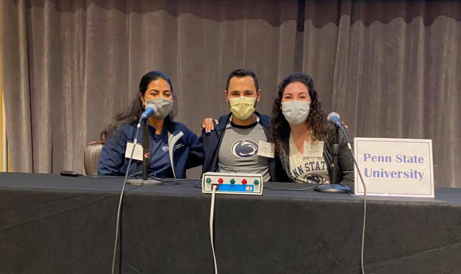 Physician Assistant students Jasmin Pimentel, Jonathan McKinney and Maria Gillio sit behind a table wearing face masks, with a sign that says Penn State University.