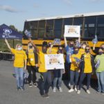 A group of 14 women stand in front of a school bus holding signs and pom-poms. They are wearing United Way T-shirts, jeans and sneakers.