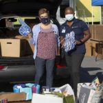 Pam Steibing, left, executive assistant, and Deborah Addo, Penn State Health chief operating officer, stand in front of a vehicle with its hatch open, showing boxes in the back. Bags of school supplies are at their feet. Pam has a foam finger on her right hand, and Deborah is holding a pom-pom shaker.