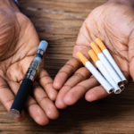 Two hands with the palms facing up hold items. The right hand contains an electronic cigarette. The left holds five cigarettes.