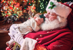 A man dressed as Santa Claus holds a sleeping infant on his chest.