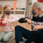 A young girl and an older man, seated next to each other on the floor near a Christmas tree, exchange elbow bumps.