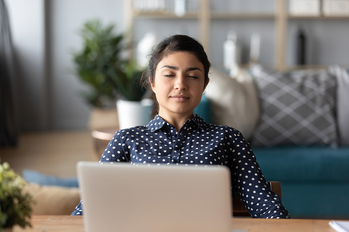 A young woman sits in front of a laptop while meditating in her living room.