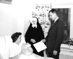 Esther Taylor, who has short hair and wears a hospital gown, lies in a hospital bed, cradling her newborn son, whose face is peeking out from a blanket. A nun and a man in a suit holding a large envelope stand by the bed.