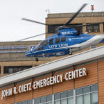 A Penn State Health Life Lion helicopter hovers above the helipad atop Holy Spirit Medical Center. A sign in the foreground on the building reads “John R. Dietz Emergency Center.” The hospital logo is in the background on the building.