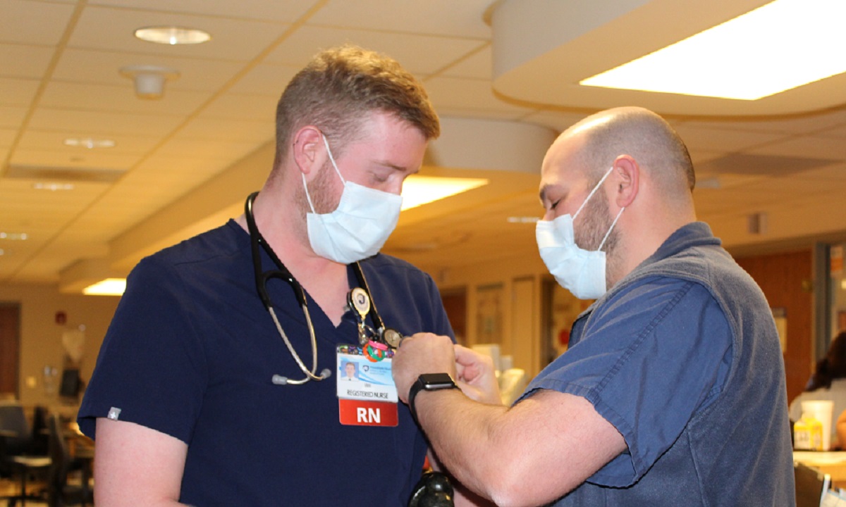 A bald man wearing a face mask pins an award on a man who has a stethoscope wrapped around his neck and is wearing a face mask.