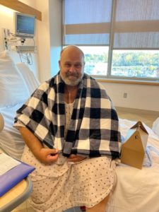 A man has a flannel blanket draped over his shoulders as he sits in a hospital bed. The man wears a gown.