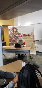A man dressed as Santa Claus sits at a desk in front of a laptop. Behind him is a screen designed to look like a mantle with a holiday tree.