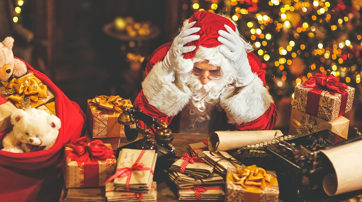 ‘Santa Claus’ rests his elbows on a desk and his head on his hands. The desk is covered with presents and letters. A Christmas tree is in the background, out of focus.
