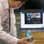 A man holds a 3D printed N95 respirator while looking at a computer screen on which several people are visible as part of a Zoom meeting.