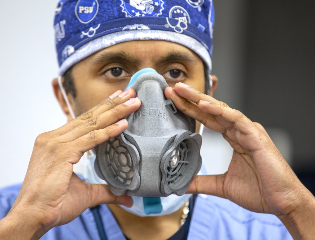 A man holds a 3D printed N95 respirator up to his face with both hands. He wears surgical scrubs and a head covering bearing Penn State athletic symbols.