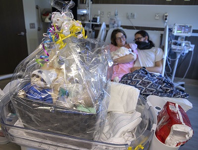A fruit basket sits on a table in a hospital room. Behind the basket, a man and a woman hold a baby while lying in a hospital bed. The man wears a mask.