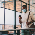 A man wearing a surgical mask walking through an office holds a cell phone in his right hand and a laptop bag over his left shoulder.
