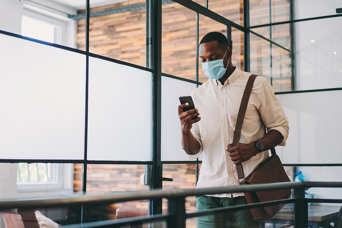 A man wearing a surgical mask walking through an office holds a cell phone in his right hand and a laptop bag over his left shoulder.