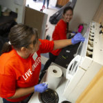 Two women wearing rubber gloves work side-by-side to clean a kitchen in a residential apartment. One looks to the other, smiling.